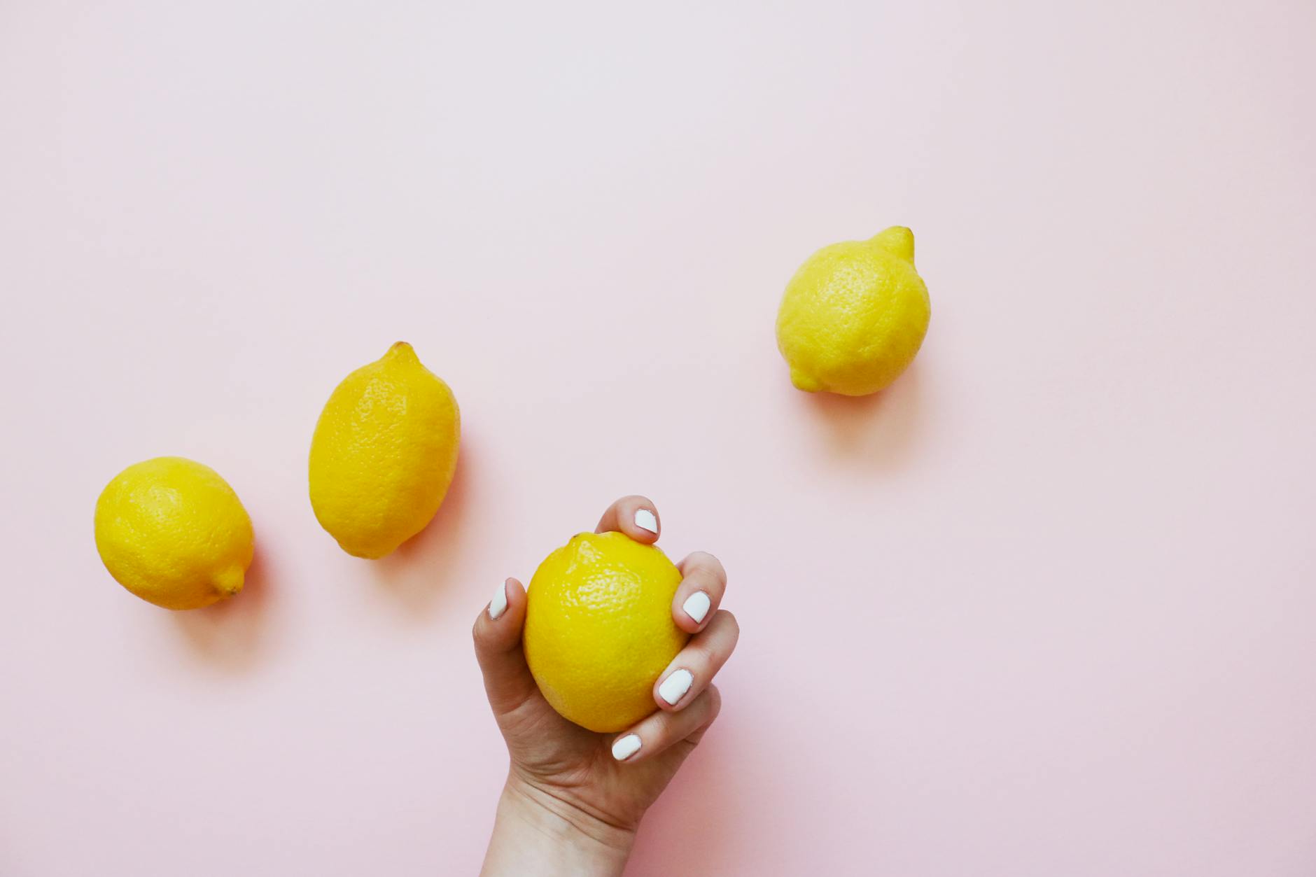 A hand holding a fresh lemon on soft pink background with additional lemons nearby, representing rest and recovery in pleasure practices