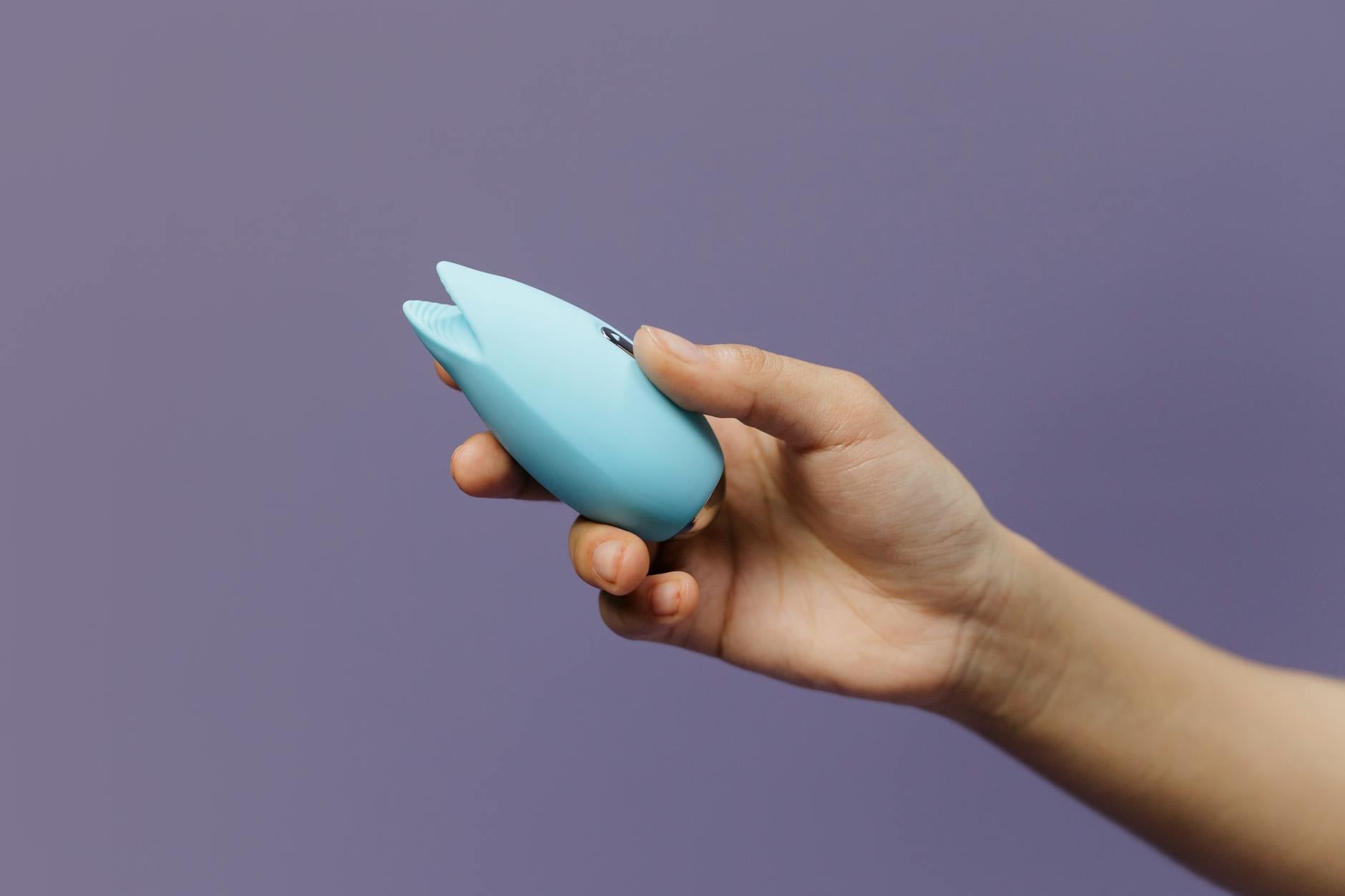 A blue silicone clitoral vibrator held in hand against a solid purple background
